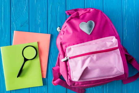 Top view of textbooks magnifier and pink backpack on blue wooden background