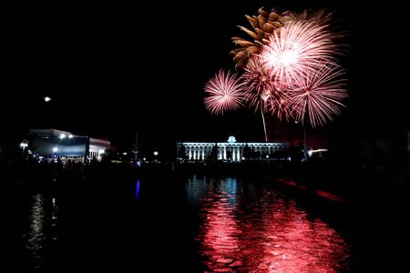 Colorful festive fireworks salute over buildings in the city and its reflection in the fountain against the black sky