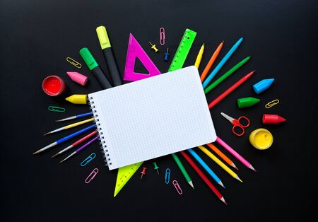 Empty checkered note pad surrounded by multicolored school supplies as fan isolated on black background with copy space