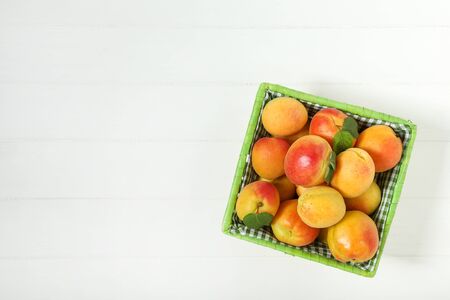 Sweet apricots with green leafs in basket on white wooden table Stock Photo