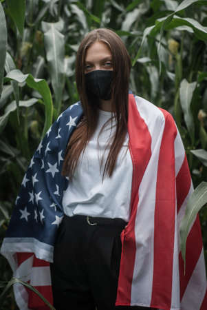 A masked girl with the usa flag on the background of a corn field labor day during the pandemic