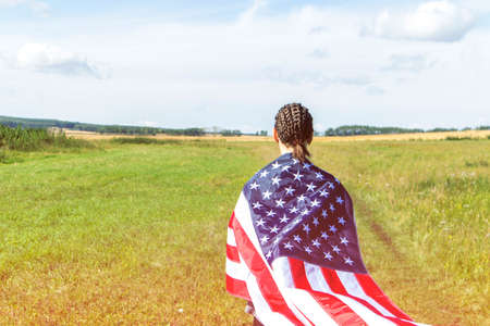 American young woman with afro braids in a field of wheat wrapped in usa flag labor day usa sunset light