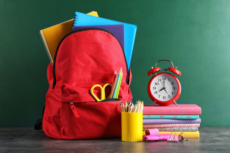 Backpack with school stationery on table against blackboard