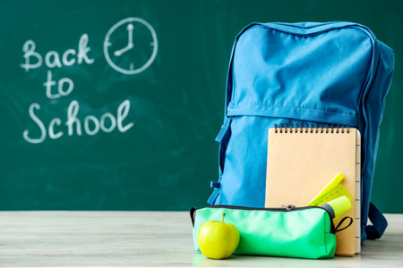 Backpack and supplies on table near green chalkboard with text back to school