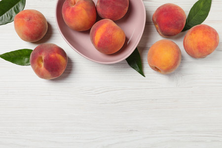 Whole many fresh ripe peaches and green leaves on white wooden table flat lay space for text