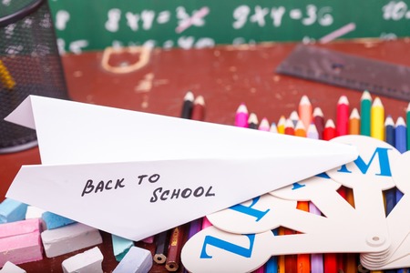 Colorful pencils of red yellow orange violet purple pink green blue paper plane with back to school text and fan english alphabet lying on brown school desk on written with white chalk blackboard