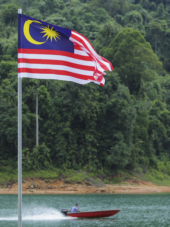 Malaysia flag jalur gemilang waving with the background of boat at the lake and malaysian rainforest trees