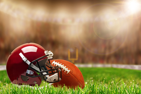 Low angle view of football helmet and ball on field grass and deliberate shallow depth of field on brightly lit stadium background with copy space