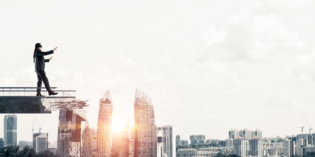 Businessman walking blindfolded on concrete bridge with huge gap as symbol of hidden threats and risks cityscape view with sunlight on background 3d rendering Stock Photo